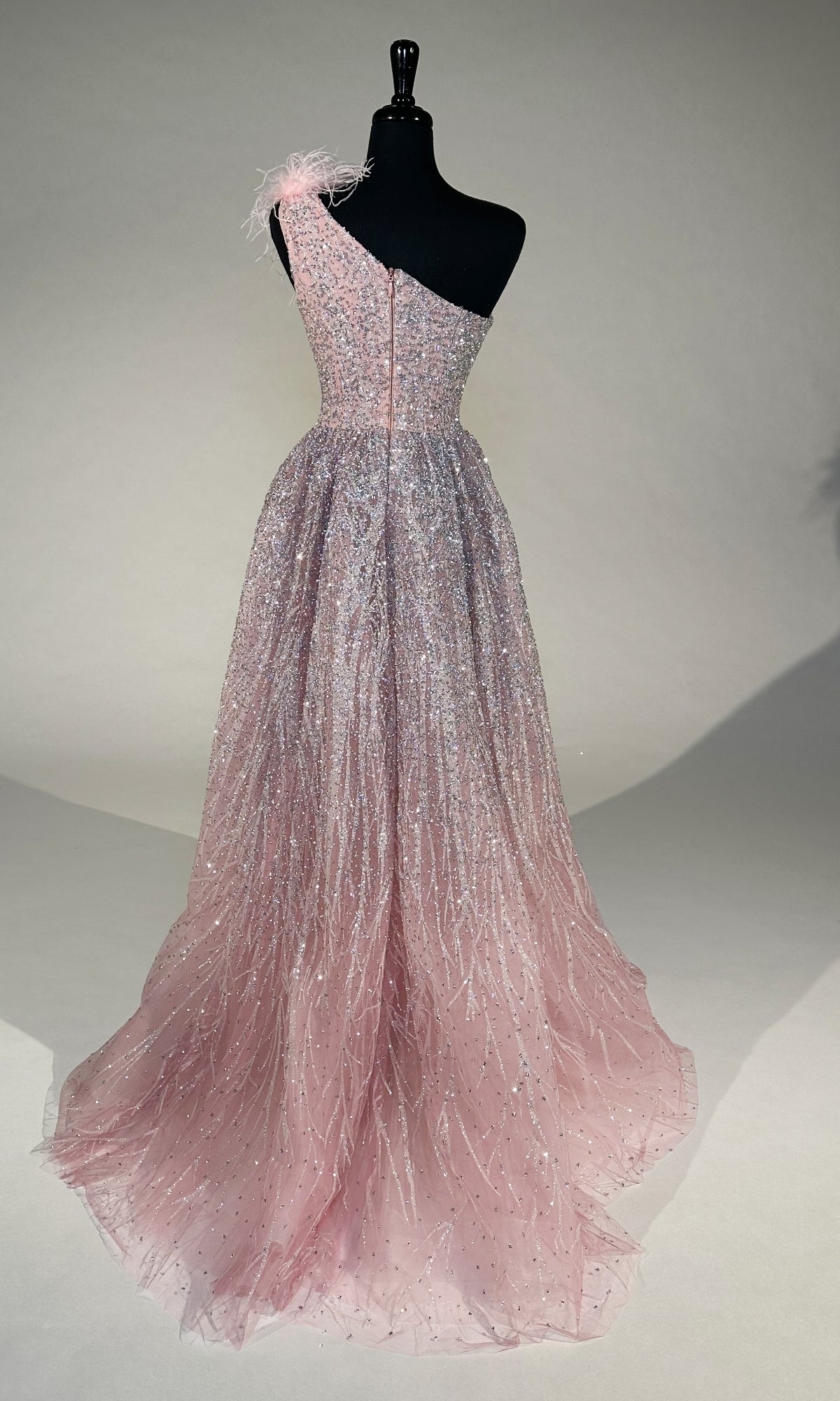 The Portia and Scarlett PS24411 Long Prom Dress is blush pink, floor-length, features one-shoulder styling with sparkling embellishments and a feather detail on the strap, shown here on a black mannequin.
